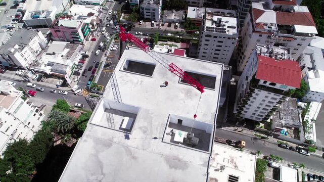 High-angle aerial drone shot orbiting left above a building under construction while a red mobile crane lowers a water tank into a rooftop opening in Santo Domingo