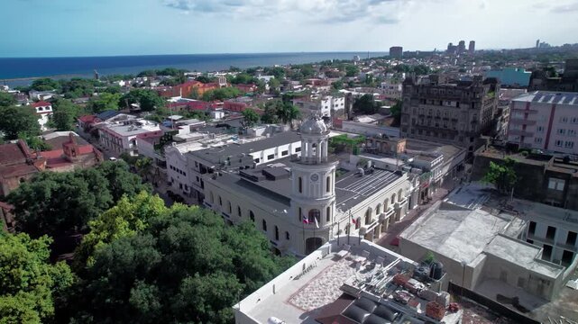 High-angle aerial drone shot advancing over Parque Colon toward the clock tower of the historic City Hall building (Palacio Consistorial) in the Colonial Zone of Santo Domingo