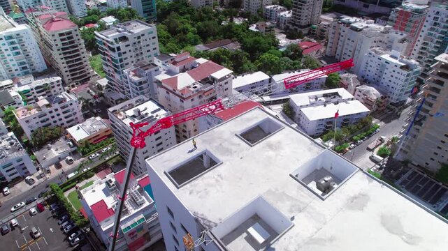 High-angle aerial drone shot advancing and ascending over a building construction site rooftop with a red telescopic crane, revealing the surrounding urban cityscape in Santo Domingo