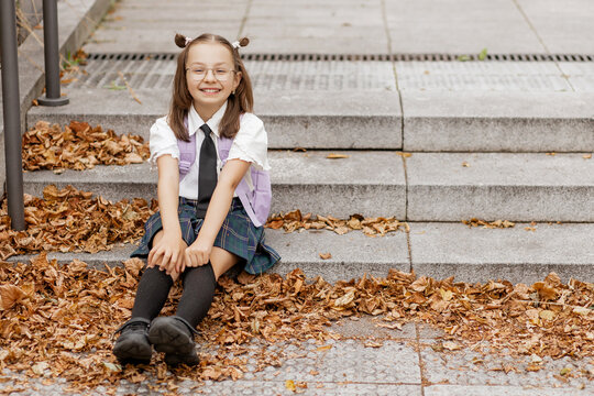 Smiling alpha generation schoolgirl with pigtails and glasses in school uniform sitting on autumn park steps outdoors. Back to school. Copy space
