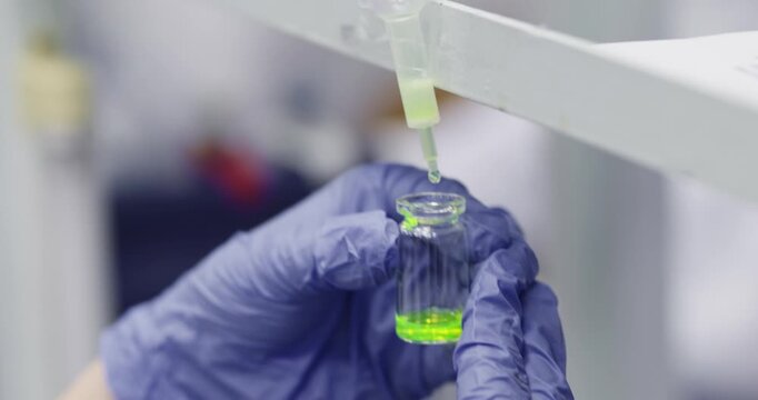 A close-up of a green glowing liquid being drop filtered using a columnar filter into a glass vial held by a scientist.