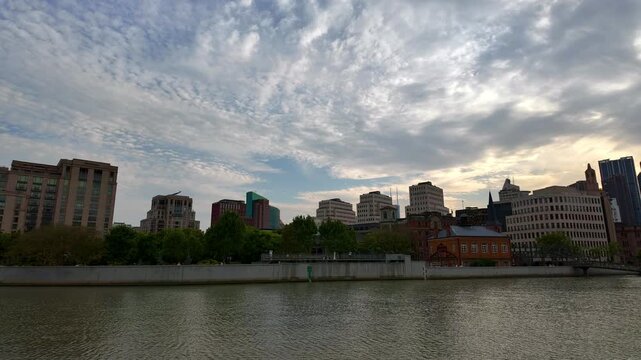 A view of an urban waterfront with various buildings, green trees, and a cloudy sky above a calm river.