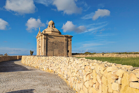 Kappella San Nikolaw chapel beside paved path and traditional Maltese stone walls, green fields and bright blue sky with scattered clouds in rural Malta