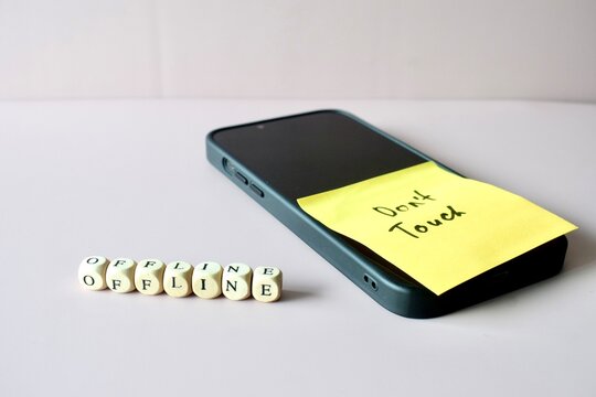 Angled shot of a smartphone with a "Don't Touch" reminder note on the screen, accompanied by wooden letter cubes spelling out "OFFLINE" on the surface.