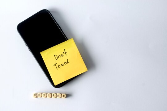 Flat lay composition of a mobile phone with a "Don't Touch" sticky note next to wooden letter beads spelling "OFFLINE" on a plain light background.