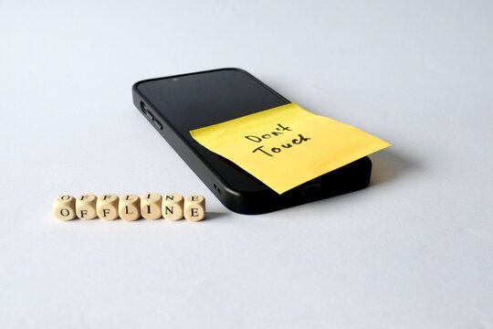 Angled shot of a smartphone with a "Don't Touch" reminder note on the screen, accompanied by wooden letter cubes spelling out "OFFLINE" on the surface.
