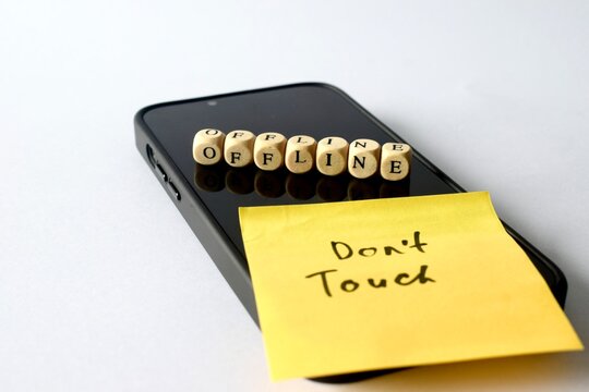 Close-up of a modern smartphone with wooden blocks spelling "OFFLINE" and a yellow sticky note reading "Don't Touch" on a white background, symbolizing a digital detox.