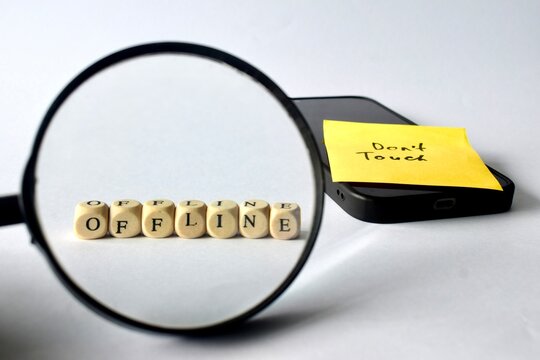 A magnifying glass focuses closely on wooden cubes spelling out the word "OFFLINE" with a smartphone and reminder note blurred in the background.