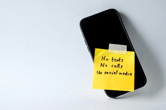 A smartphone lies flat on a white surface with a yellow note taped to the screen reading "No texts, No calls, No social media," showcasing a digital detox concept.