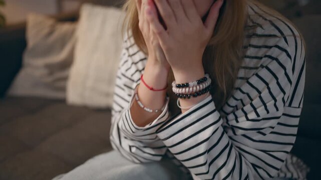 Upset teenage girl sitting on sofa and covering face with hands, showing emotional distress caused by bullying, school problems, social pressure and difficult adolescence.