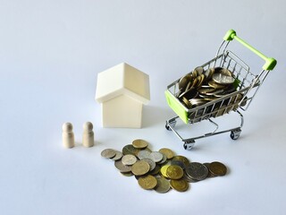 White toy house, two standing wooden peg dolls, a heap of coins, and a miniature shopping cart overflowing with money, conceptualizing family home purchases and real estate savings. © Akram