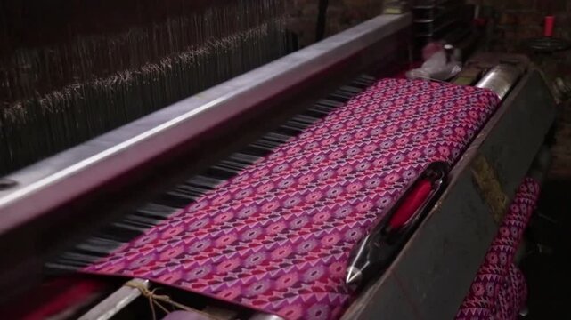 A modern automatic mechanical loom in operation, weaving a fabric with a geometric pattern dominated by pink and purple.