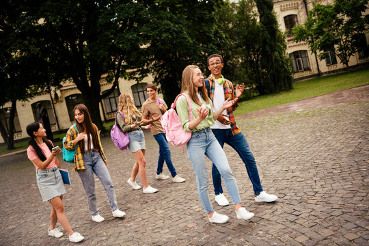 Group of happy college students walking and talking together on a leafy campus courtyard, laughing and carrying backpacks in casual spring clothing