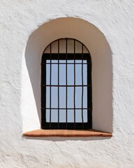 Fototapeta premium Arched iron grilled window set in white plaster wall with clear sky reflection for architectural detail, texture background, facade study and minimal design projects