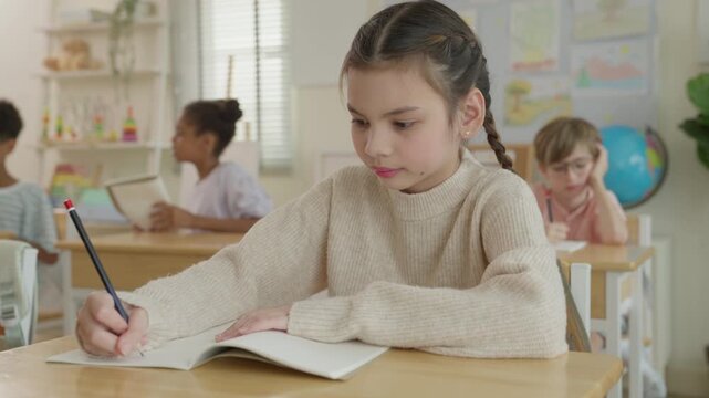 Girl writing in notebook at classroom desk with classmate, studying focused expression during school lesson, colorful educational materials and teacher nearby in background