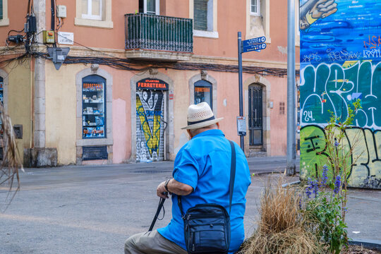 Senior Man Sitting on Street in Barceloneta Neighborhood, Barcelona, Blue Shirt and Straw Hat Back View, Traditional Shop Facade
