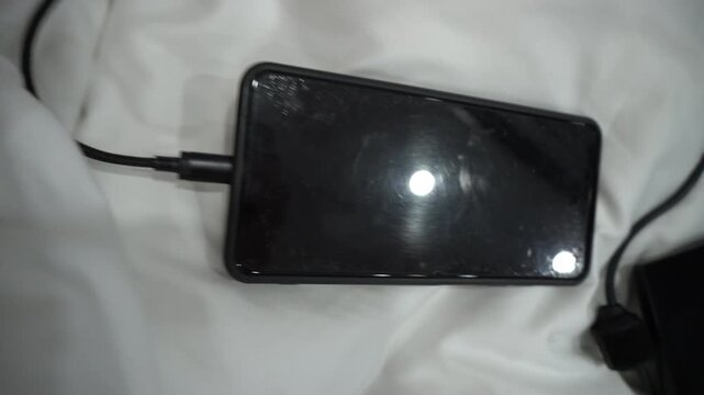 Black smartphone charging on a white bed sheet with visible cable  