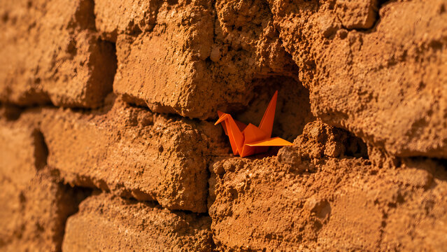 Vibrant orange origami paper crane placed inside a gap of a rustic mud brick wall