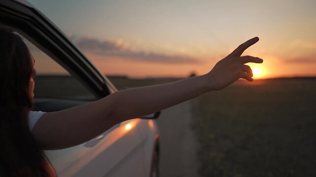 Girl reaches hand to touch sun on summer road. Freedom shines through car window. Sunset glows over travel adventure. Girl moves hand in wind. Car on road to summer freedom with light and adventure