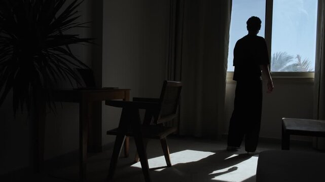 Silhouette of lonely man slowly walking through dark room and stopping by window, expressing solitude, emotional isolation, sadness, emptiness and quiet despair