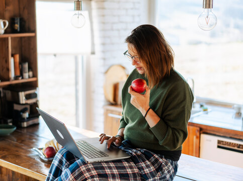 Happy middle-aged woman sitting on table with red apple, looking at laptop screen. Healthy lifestyle at work.