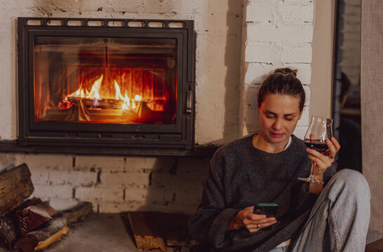 Cozy winter evening at home. Beautiful young woman sitting near fireplace with glass of wine and smartphone.