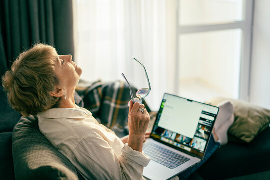 Senior Caucasian woman leaning back on a sofa and relaxing with a closed laptop on her knees.