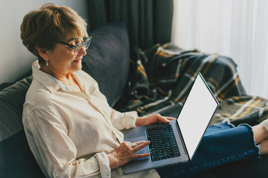 Happy senior Caucasian woman in glasses sitting on a sofa with a laptop on her knees, white isolated screen.