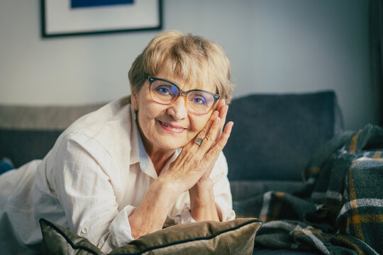 Cheerful senior Caucasian woman lying on her stomach on a sofa, propping her head with hands and smiling at the camera.