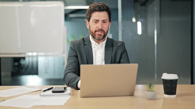 Focused professional working in modern office environment. Man engages with laptop, making video call, enjoying coffee break. Demonstrates productivity and remote work business communication methods.