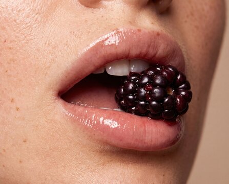 Glossy lips biting ripe blackberry close up with natural skin texture and white teeth
