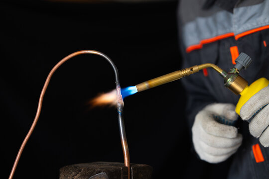 Close-up of a worker in protective gloves brazing a copper pipe with a gas torch. Professional plumber soldering thin copper tubing with a hot blue flame in a workshop, plumbing and HVAC installation 