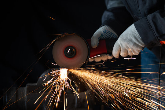 a worker in protective gloves cutting a metal bar with an angle grinder tool in a workshop. Stream of bright orange sparks flying from the abrasive disc against a dark background.