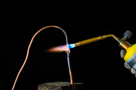 Close-up of a propane gas torch heating a copper pipe during a brazing or soldering process. Professional plumbing or HVAC installation with a hot blue and orange flame against a dark background