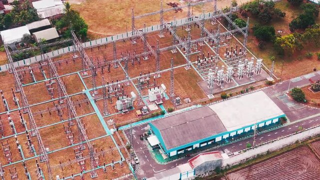 Aerial drone view of a large electrical substation featuring high voltage transformers, transmission towers, steel structures, switchgear, and power distribution equipment surrounded by dense urban bu
