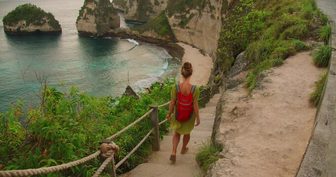 Woman tourist walks down steep stone stairs to tropical coastline toward Dimond beach in Nusa Penida Island Bali