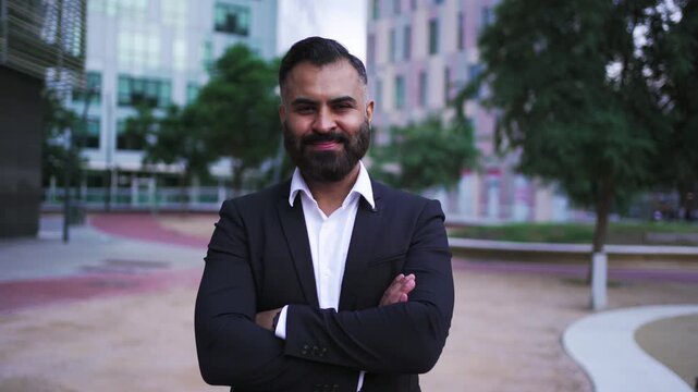 Confident bearded Indian man standing in a modern city business district, looking at camera cheerfully.