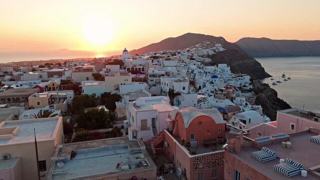 Warm sunrise light washes over Oia&rsquo;s tiered rooftops and rugged coastline as the drone glides above the cliff town and sea.