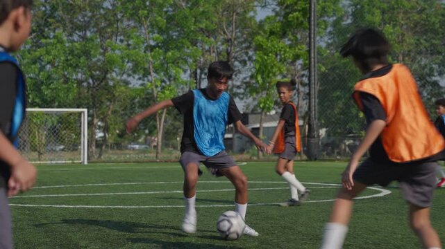 Youth Soccer Player Dribbling Ball Across Outdoor Artificial Turf Field as Teammates Defend and Practice Teamwork Near Green Trees and Chain Link Sports Fence
