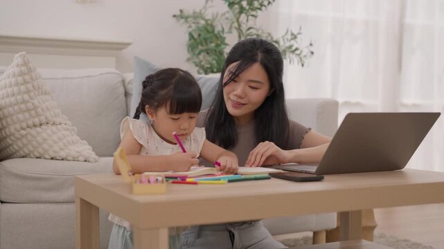 Asian mother and daughter drawing home table beside laptop, creating colorful pencil art and homework together, warm family learning activity with focused child, woman, paper materials, and creative