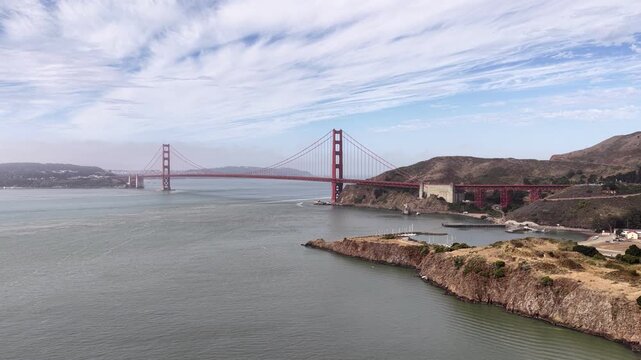 Aerial drone footage from Sausalito, California, captures the famous Golden Gate Bridge spanning San Francisco Bay, framed by the Marin Headlands, coastal hills, and expansive blue waters