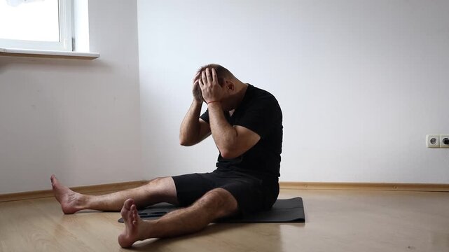 Man sitting on yoga mat performing seated forward bend to stretch his hamstrings, exercising at home in bright empty room
