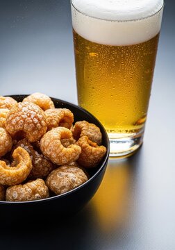 Close up of crunchy golden pork cracklings served in a minimalist black bowl beside a cold glass of refreshing beer with froth and bubbles, nibbles, savory, cold