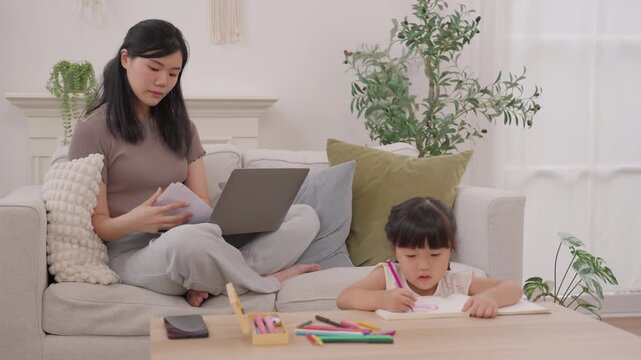Asian mother helping child with drawing and laptop at home, family activity on cozy sofa, young woman supporting daughter with colorful pencils, warm indoor lifestyle scene and creative learning time