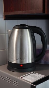 Stainless steel electric kettle with black handle and red power indicator light, standing on a kitchen shelf beneath wooden countertop