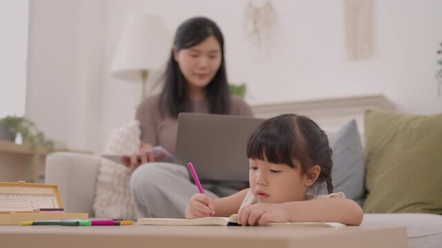 Asian mother and daughter study education together with laptop and coloring, while woman and child enjoy focused family learning activity at home in bright living room with creative expressions