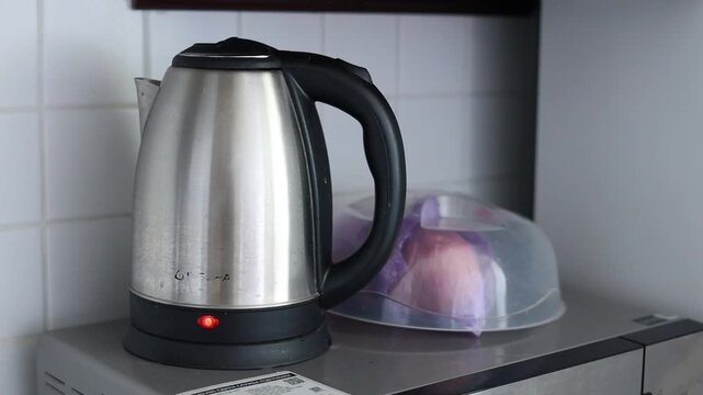 Stainless steel electric kettle with black handle and red power indicator light, standing on a kitchen shelf beneath wooden countertop