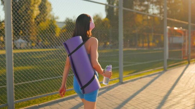 Rear view of a fit woman carrying a yoga mat along the sunny park fence.