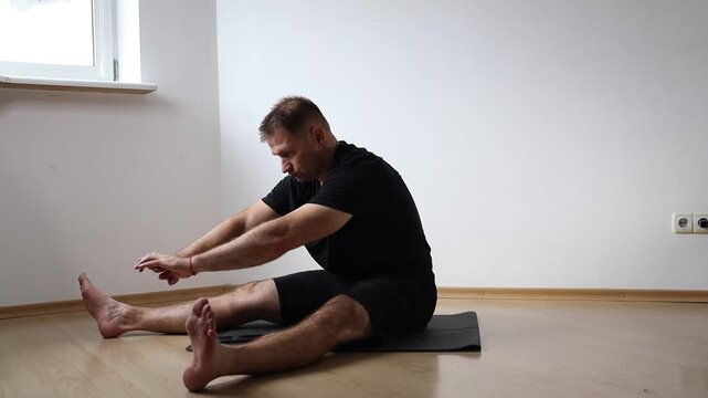 Man sitting on yoga mat performing seated forward bend to stretch his hamstrings, exercising at home in bright empty room