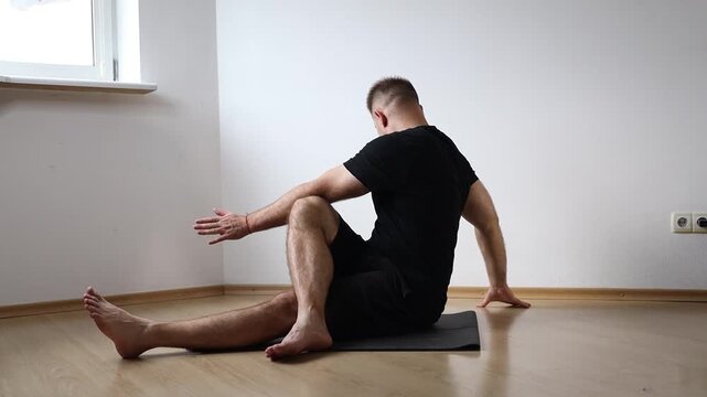 Man sitting on yoga mat performing seated forward bend to stretch his hamstrings, exercising at home in bright empty room
